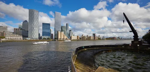This urban landscape photograph, taken in the afternoon during early autumn, features Canary Wharf Dock Crane 1 prominently situated along the River Thames in London, United Kingdom. The image captures the historic dockside crane to the right, with the modern skyline of Canary Wharf rising across the water. Landmark skyscrapers, including the HSBC building and distinctive contemporary towers, are clearly visible in the background, illustrating the business district's architectural diversity. The river itself is central to the composition, with boats moving across its surface under a sky filled with clouds, encapsulating the dynamic urban environment of this area.